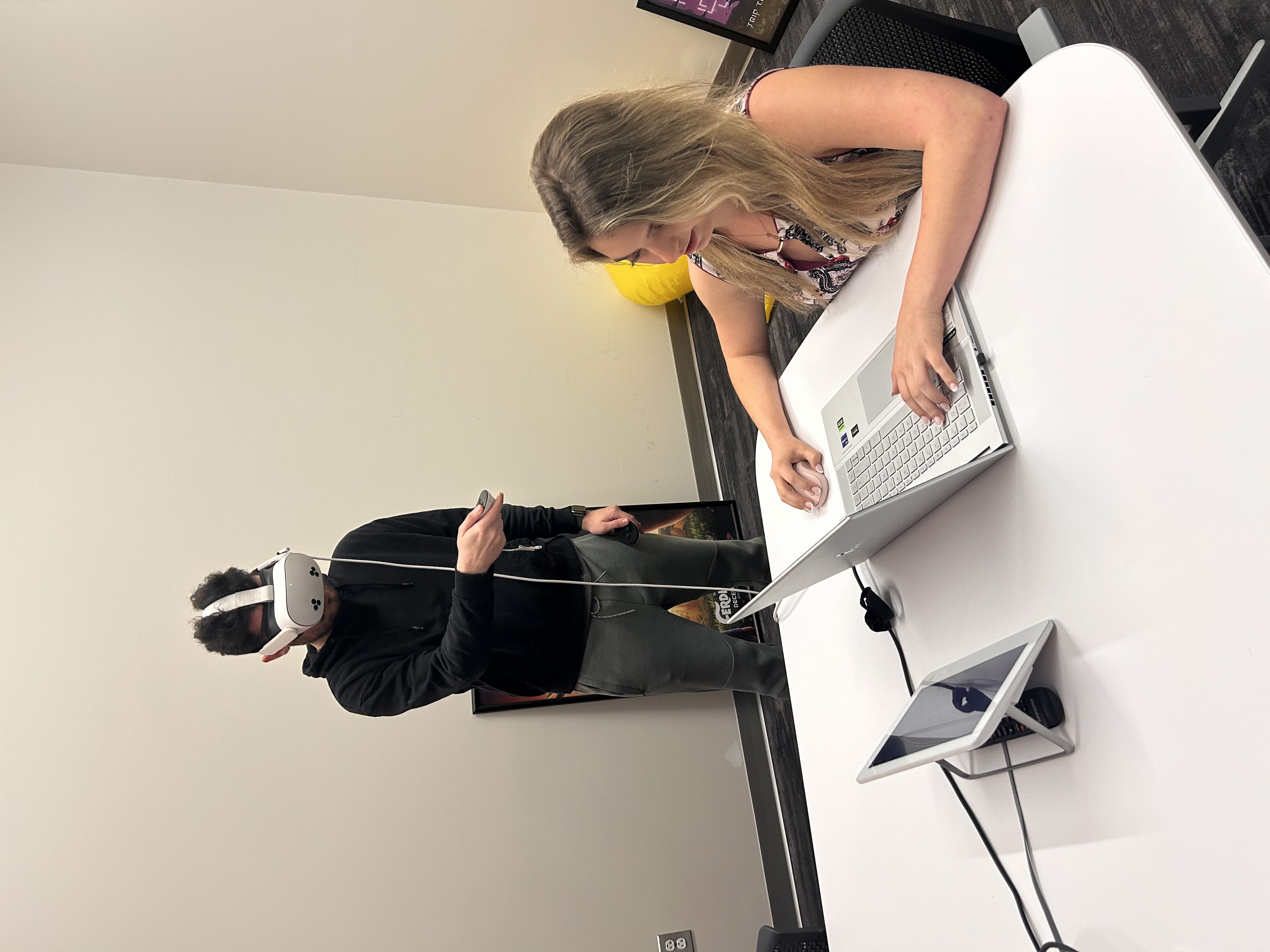 One person is standing up and wearing a VR headset and the other person is using a laptop with a mouse at a table
